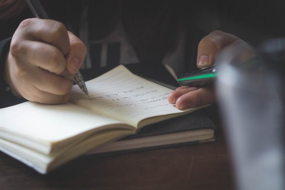 Mão escrevendo em um caderno aberto sobre a mesa, em ambiente com pouca luz