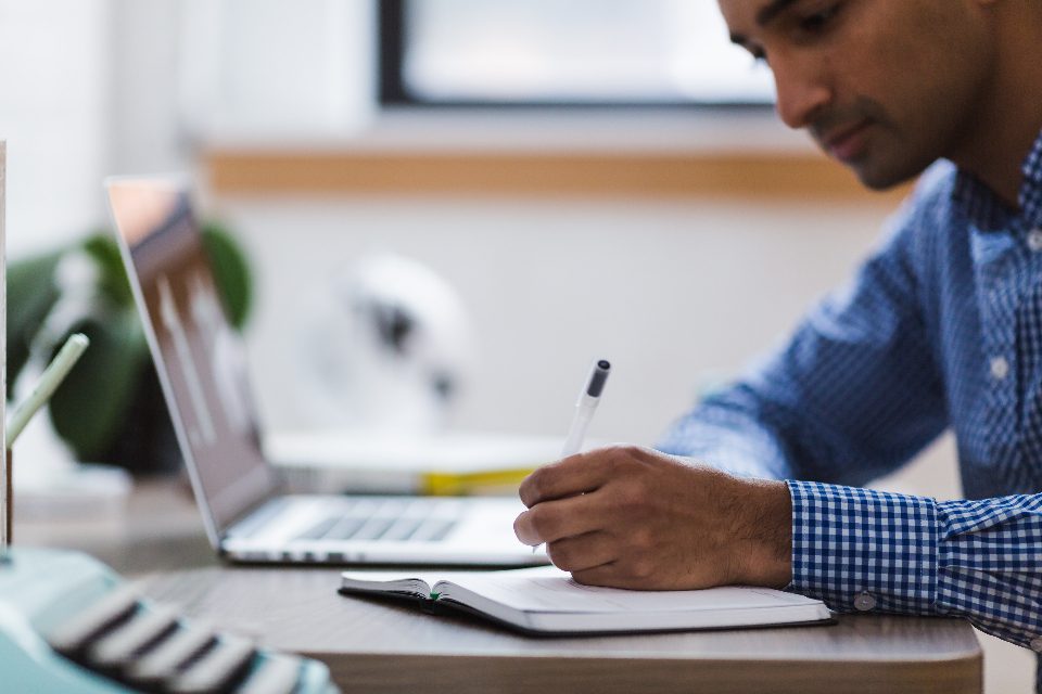 Homem anotando em um caderno ao lado do notebook enquanto começa uma tarefa