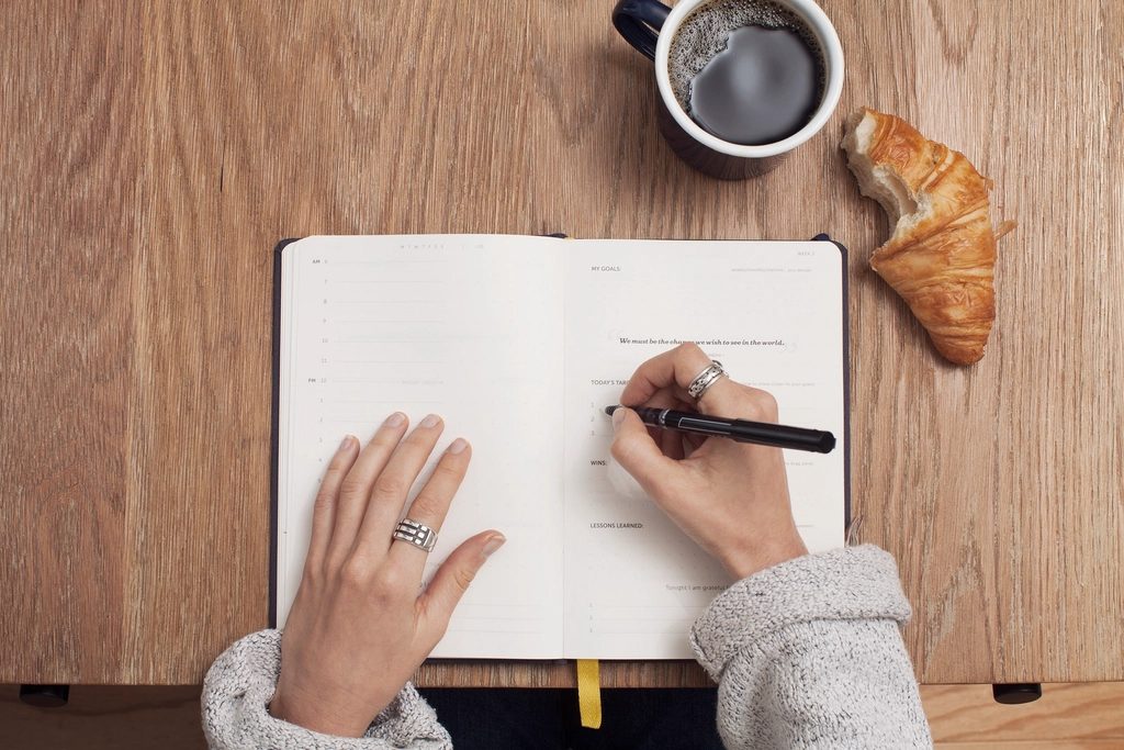 Mãos escrevendo em uma agenda semanal ao lado de uma xícara de café